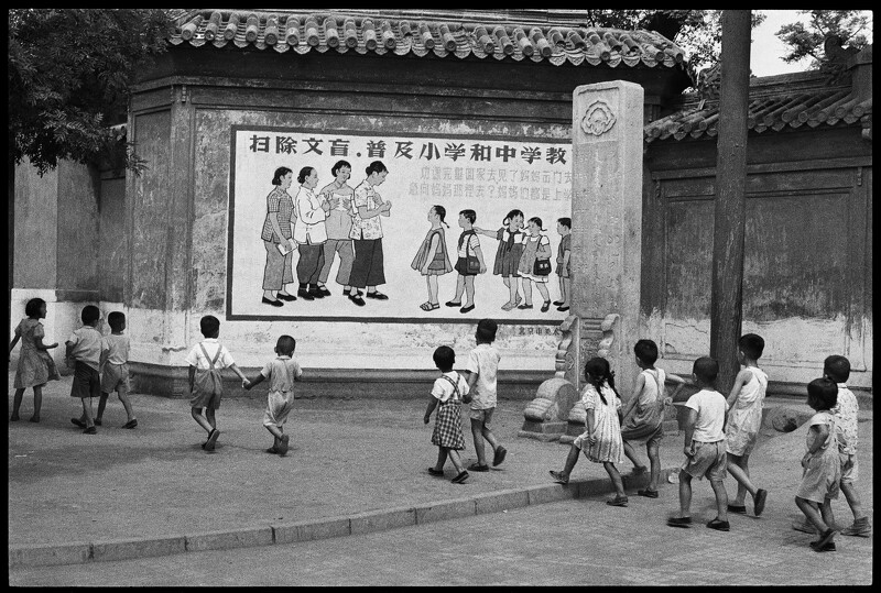 《孩童正從一幅寫著「…媽媽們都是上學去…」宣傳標語的看板前經過,北京,1958年7月》