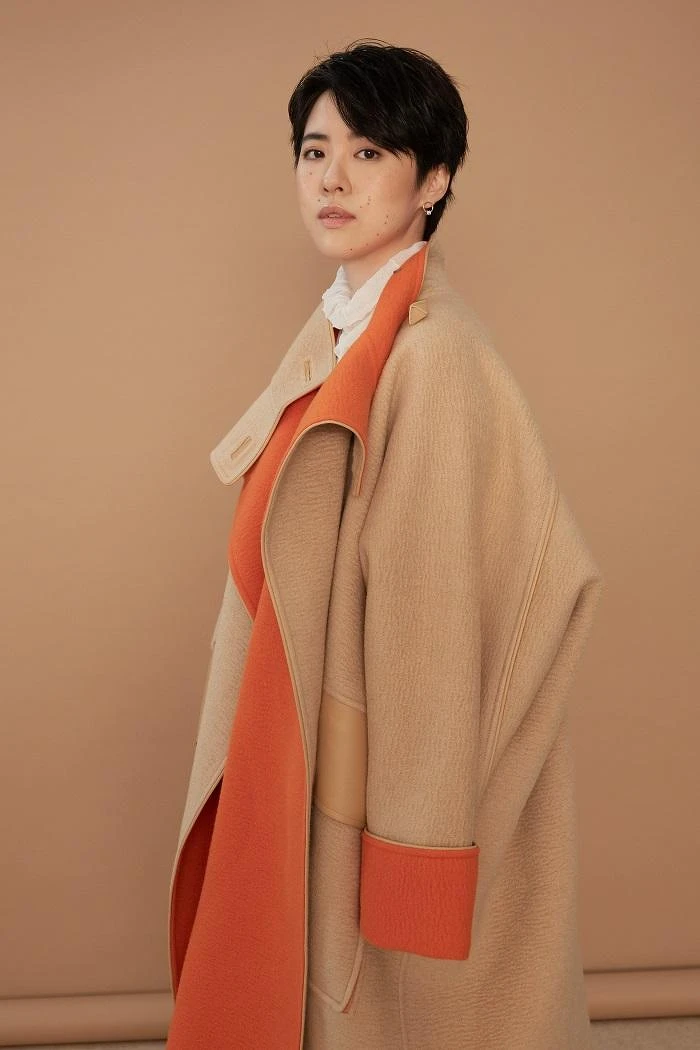 白高領透膚上衣 LOEWE；雙色駱駝斗篷大衣、米色馬褲、小牛皮長靴All by HERMÈS；幾何金耳扣、圓圈線條耳扣、圈形金耳環、字母線條戒指、金色戒指All by APM MONACO。