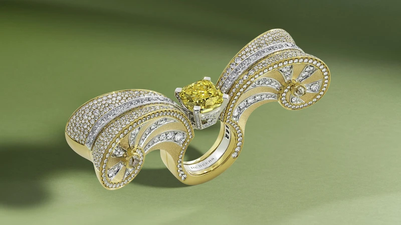 Metamorphosis by De Beers 系列 Summer Masterpiece 鑽石與嵌套式戒指，主鑽為重達7.61 克拉的艷彩黃鑽，戒指外套部分雕塑感的精緻螺旋形狀由磨砂和拋光18K黃金巧製，以鑽石鑲嵌出盤繞線條，並以黃色鑽石原石作結，猶如砂礫中隱藏的珍貴化石。